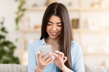 Harika bir uygulama. Neşeli genç Asyalı kadın evde modern akıllı telefona bakıyor, sosyal ağları kontrol ediyor ya da internetten alışveriş yapıyor, haberleri okuyor, izin gününün tadını çıkarıyor, oturma odasında bir aletle koltukta oturuyor.