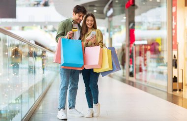 Mobil Alışveriş. Bir çift alıcı telefon ve kredi kartı kullanıyor. Uygulamada yeni kıyafetler alıyorlar. Renkli alışveriş torbaları modern alışveriş merkezinde duruyor. Ticari Teklif ve Satış