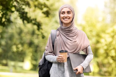 Mutlu Müslüman iş kadını parkta yürüyor, kahve içiyor, dizüstü bilgisayarı tutuyor, boş bir yer tutuyor.