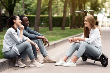 Üç farklı genç, bir şehir parkında kahkahalar ve hikayeler paylaşıyor. Biri motosikletle otururken, diğerleri yakınlarda dinleniyor. Güneş ağaçların arasından parlıyor, canlı bir atmosfer yaratıyor..