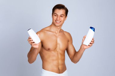Kozmetik Ürün Şablonu. Tişörtsüz bir adam gri stüdyo duvarından izole edilmiş şampuan ve duş jeli tüpleri tutuyor.