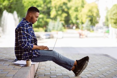 Sınav Hazırlığı. Siyahi bir öğrencinin, elinde kahve ve defterlerle parka otururken, bilgisayarını kullanmasının tam boy portresi.