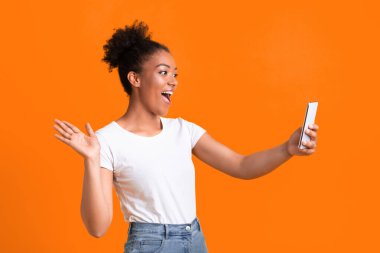 Bağlantı. Heyecanlı Afrikalı kız telefonla video görüşmesi yapıyor ve arayana el sallıyor, Turuncu Stüdyo Duvarı, bedava alan