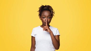 Sessiz siyah bir kadın sessiz bir jest yapıyor. Fotokopi alanı olan sarı bir stüdyo. Çekici Afrikalı Amerikalı genç bayan sessiz bir jest yapıyor, tek parmağıyla dudaklarına dokunuyor.