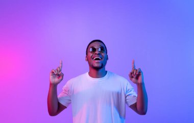 Neon ışıklarının altında dikilen heyecanlı siyahi adam fotokopi uzayını işaret eden, şık güneş gözlüklü neşeli Afrikalı Amerikalı adam, Mor Arkaplan üzerinde boş yer gösteriyor