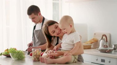 Neşeli bir aile evde yemek hazırlığı yapıyor. Gülümseyen anne bebeğiyle oynarken baba sebzeleri doğruyor ve sıcak ve sevgi dolu bir ortam yaratıyor..
