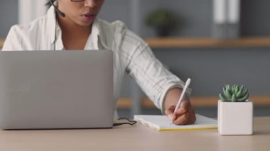 Bir kadın, dizüstü bilgisayar ve not defteri kullanarak evden ders veriyor. Modern online eğitim teknikleri sergileyerek öğrencilerle uzaktan iletişim kuruyor..