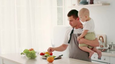 Bir baba, bebeğini tek koluyla tutar. Güneşli bir mutfakta taze meyve hazırlarken gülümser. Oda, tezgahında yeşil marul ve renkli meyvelerle parlak bir tasarım içeriyor..