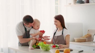 Ebeveynler yemek hazırlıklarına yardım ederken bir çocuk mutfak tezgahına oturur. Taze sebzeler yayılıyor ve parlak, havadar alanda sıcak, sevgi dolu bir atmosfer yaratıyor..