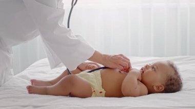 İlgili bir tıp uzmanı, bir çocuğu evde muayene eder. Çocuk yatakta rahat bir şekilde yatıyor, kontrol sırasında merak ve sakinlik gösteriyor, güven ortamı yaratıyor..