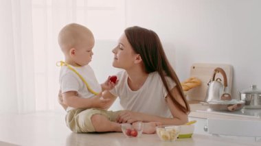 Yeni yürümeye başlayan bir çocuk mutfak tezgahında oturup annemle bir şeyler atıştırıyor. Atmosfer sıcak ve parlak, yemek saatinde sevgi ve özenle dolu rahat bir ev ortamı sergiliyor..