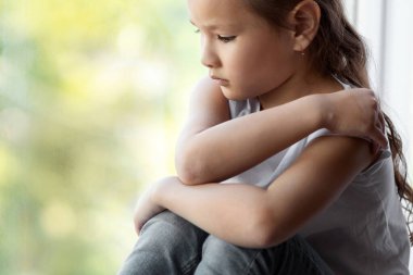 Çocukluktaki depresyon. Pencerenin yanında oturan mutsuz çocuk yalnızlıktan acı çekiyor ve kendini evinde kucaklıyor. Metin için Boş Alan