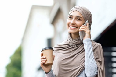 Alış veriş yapan neşeli Müslüman kadın sokakta akıllı telefondan konuşuyor, kız arkadaşlarına mevsimlik satışlardan bahsediyor, kahve içiyor, fotokopi çekiyor.