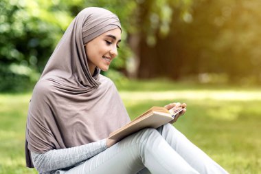 Rahat Müslüman kadın parkta eğleniyor, çimenlerde oturuyor ve kitap okuyor, boş bir alan.