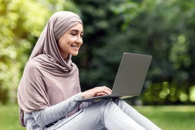 Kamu parkında dinlenirken dizüstü bilgisayar kullanan güzel müslüman kızın yan görüntüsü, boş alan