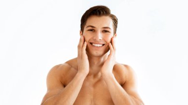 Erkek Cilt Bakımı. Çekici kaslı adamın ön portresi çıplak gövdesi yüzüne dokunuyor ve gülümsüyor, beyaz arka planda soyutlanıyor. Stüdyoda poz veren gömleksiz adam, kameraya bakıyor.