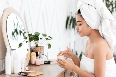 Havluya sarılı genç Asyalı kadın evde sabah yüz kremi kullanıyor, yan görüş, fotokopi odası. Gülümseyen kadının elinde besleyici ya da nemlendirici yüz kremi olan bir kavanoz var.