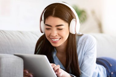 En sevdiği eğlence yeri. Genç Asyalı kız dijital tablet ve kablosuz kulaklıkla evdeki koltukta dinleniyor, internete bakıyor ya da müzik dinliyor.