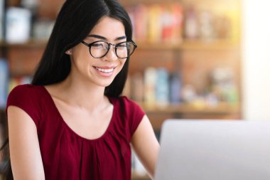 Çevrimiçi eğitim kavramı. Güzel Asyalı kız kafede dizüstü bilgisayarda çalışıyor, gözlük takıyor ve kahve içiyor, boş yer var.