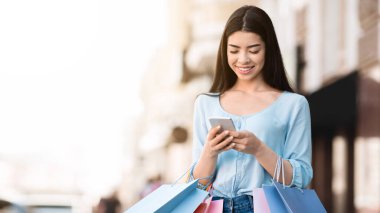 Alışveriş merkezinde başarılı bir alışveriş yaptıktan sonra akıllı telefonu açık havada kullanan genç Asyalı kız, nakit para veya kredi kartı bakiyesini kontrol ediyor