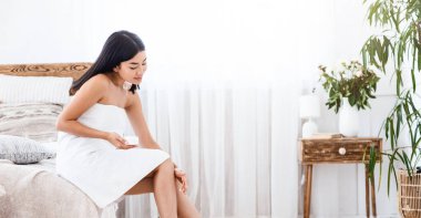 Duştan sonra güzel bacaklarına nemlendirici süren seksi Asyalı kız. Yatak odası iç mekan, yan manzara, serbest alan.