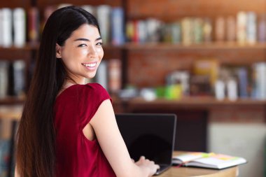 Uzaktan İş Bulma. Kahvesinde siyah ekranlı dizüstü bilgisayar kullanan, gülümseyen Asyalı genç kadın, kameraya bakıyor,