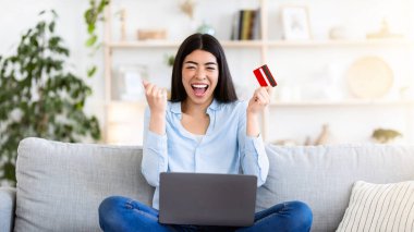 Online Credit Concept. Neşeli Asyalı kız, başarıyı banka kartı ve dizüstü bilgisayarla kutluyor koltukta oturuyor, iyi haberlere duygusal tepki veriyor.