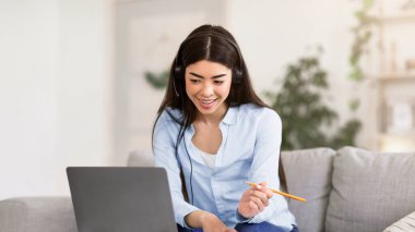 Çevrimiçi Eğitim. Kulaklıklı genç Asyalı kız evde dizüstü bilgisayarla ders çalışıyor, Webinar 'ı izlerken not alıyor.