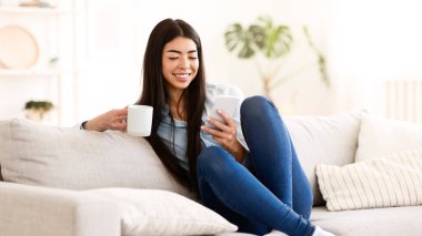 Haftasonu rahatla. Mutlu Asyalı kız akıllı telefon kullanıyor ve sabah kahvesini yudumlarken evde oturuyor oturma odasında dinleniyor.