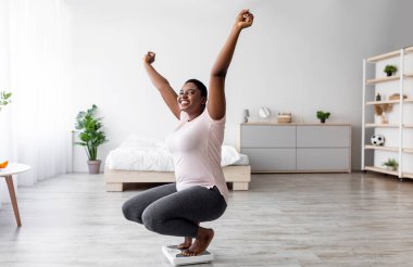 Başarılı kayıp konsepti. Aşırı neşeli, tombul siyah kadın terazide oturuyor, heyecan içinde ellerini kaldırıyor, zayıflama hedefine ulaşıyor, başarısını kutluyor, tam boy. Boşluğu kopyala