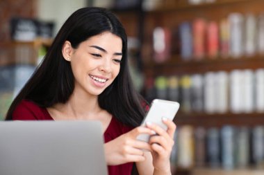 Gülümseyen Asyalı blogcu kız kafede akıllı telefon kullanıyor, yeni içerik yüklüyor, sosyal ağlardaki paylaşımı paylaşıyor, boşluğu kopyalıyor