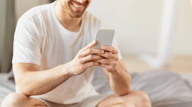 Evdeki yatak odasında yatağında oturan cep telefonu uygulaması olan tanınmayan adam cep telefonuna odaklanıyor. Smartphone 'da mesaj çeken adam. Kırpılmış, Seçici Odaklanma