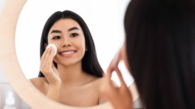 Gülümseyen Asyalı kadın yüzünü temizliyor, pamuk pedlerle ve temizlik ürünleriyle aynaya bakıyor. Genç ve çekici bir bayan yüz toneri ve pamuk pedi kullanıyor. Ev içi, boş alan.