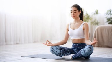 Yaşam Uyumu. Sakin Asyalı kız evde nilüfer pozisyonunda meditasyon yapıyor, oturma odasında yoga yapıyor, boşluğu kopyalıyor.