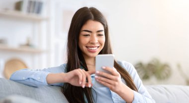 Hafta sonu eğlencesi. Gülümseyen Asyalı kız internette akıllı telefonuyla geziniyor, oturma odasında kanepede dinleniyor, boş yer var.