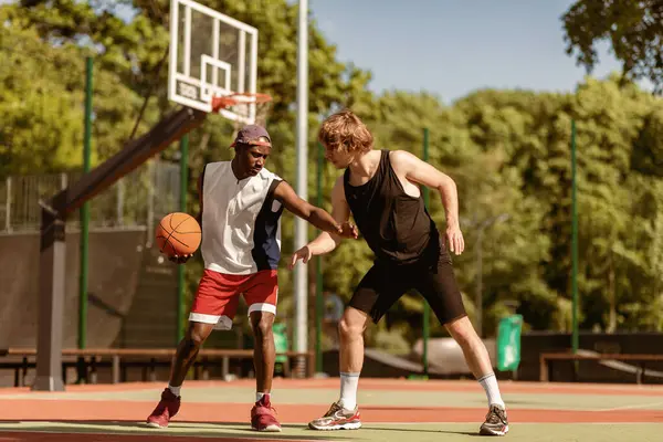 Açık hava sahasında iki profesyonel basketbolcu antrenman yapıyor. Afrika kökenli Amerikalı ve Kafkasyalı sporcular dışarıda top oynuyorlar, birlikte oyun oynuyorlar.