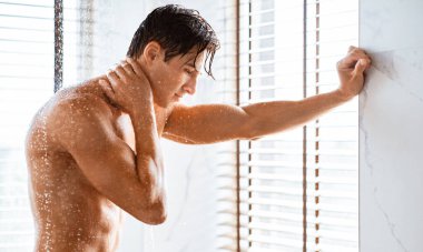 Dalgın kaslı adamın banyoda düşen suyun altında duvara yaslanıp boynuna dokunuşunun yan görüntüsü.