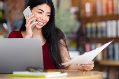 Uzak İş Konsepti. Genç Asyalı iş kadını Kafe 'de dizüstü bilgisayarla çalışırken belgelere bakıyor ve cep telefonuyla konuşuyor.