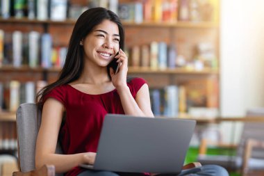 Uzaktan bilgisayarla çalışan, kahve dükkanında cep telefonuyla konuşan, rahat koltukta oturan, gülümseyen Asyalı kadın.