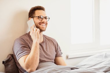 Cep telefonuyla konuşan bir adam evde yatağında oturan bir telefon görüşmesi yapıyor. Yatak odasında akıllı telefondan konuşan adam. Mobil İletişim Konsepti. Boş Alan