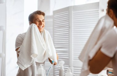 Sabah Rutini Konsepti. Gülümseyen yakışıklı genç adam modern banyoda dururken yüzünü havluyla siliyor. Kendine güvenen adam aynada kendine bakıyor, omzunun üstünden bakıyor.