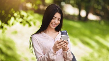 Sırt çantalı neşeli Asyalı genç kız dışarıda akıllı telefon kullanıyor, sosyal medyada geziniyor, kampüste dinleniyor.