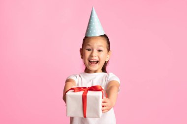Tatiller için tebrikler. Beyaz tişörtlü, parti şapkalı, kırmızı kurdeleli, pembe stüdyo arka planında izole bir şekilde duran Asyalı kızın portresi.
