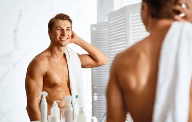 Sabah Rutini Konsepti. Tişörtsüz, gülümseyen, saçlarını eliyle tarayan, duştan sonra aynaya bakan, banyoda dikilip, omzunun üstünden bakan,