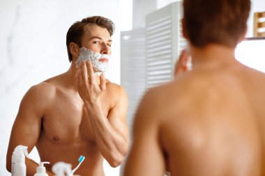 Yakışıklı, gömleksiz adam yüzüne krem sürüyor, sakalını tıraş ediyor ve aynaya bakıyor, portre yapıyor.