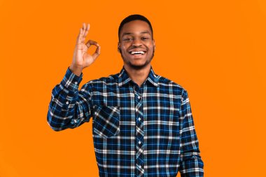 Her şey yolunda. Neşeli siyahi adam el kol hareketi yapıyor. Stüdyoda duran kameraya gülümsüyor. Sarı arka plan üzerinde.