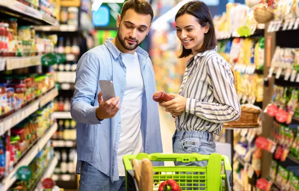 Süpermarkette alışveriş yapan genç bir ailenin portresi. Gülümseyen Çift Kapalı Kapılar 'da Gıda Ürünleri Taraması Yapıyor. Müşteriler Malzeme Alırlar, Fiyatı Karşılaştırırlar