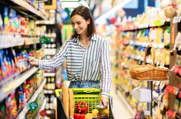 Satış, Alışveriş, Tüketim ve İnsanlar Konsepti. Gülümseyen genç bir kadın, elinde bir sepet dolusu ekolojik ürünle markette rafların yanında yürüyor, markette sağlıklı yiyecekleri seçiyor.