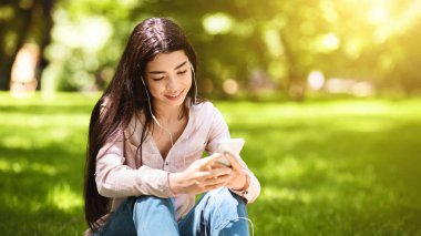 Hayalperest Asyalı kız dışarıda akıllı telefon ve kulaklıklarla en sevdiği şarkıları dinliyor çimlerin üzerinde oturmuş, müzik dinliyor.