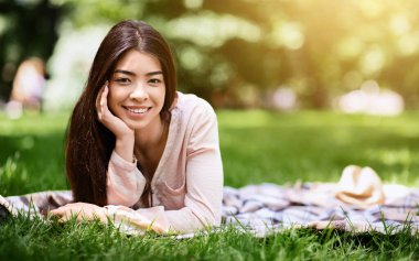 Parkta ekose üzerinde dinlenen, çimlerin üzerinde uzanan ve kameraya gülümseyen sevimli Asyalı kız.